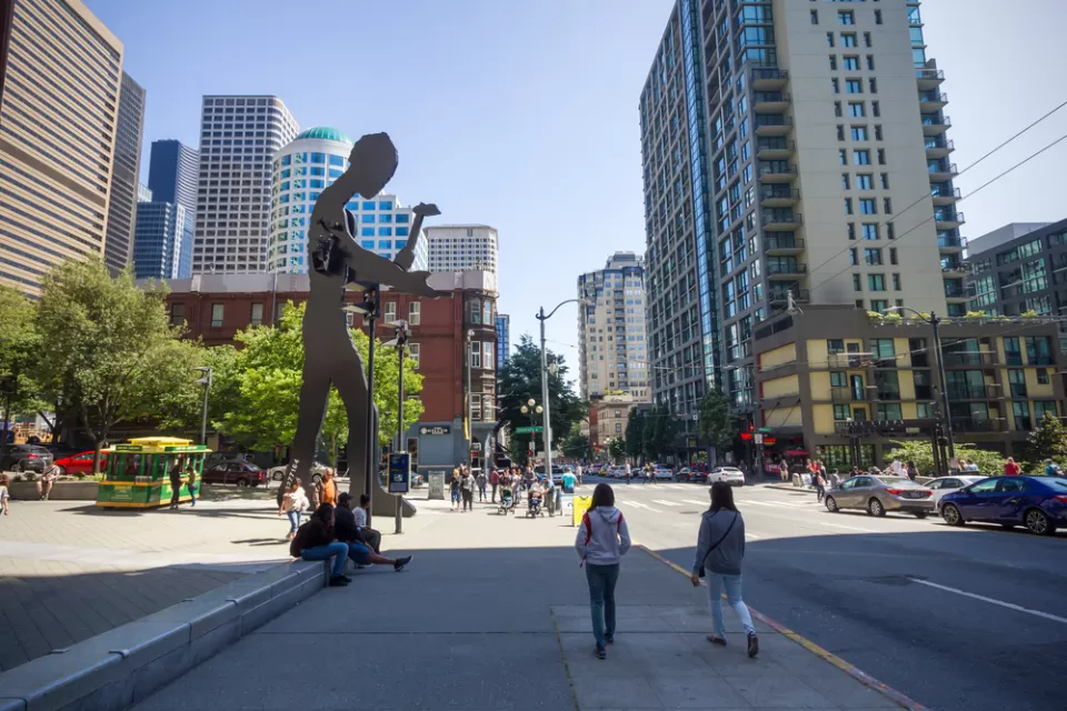 People walking on a sidewalk in front of the Seattle Art Museum, which have the hammering man and a skyscraper in the background, Washington, USA